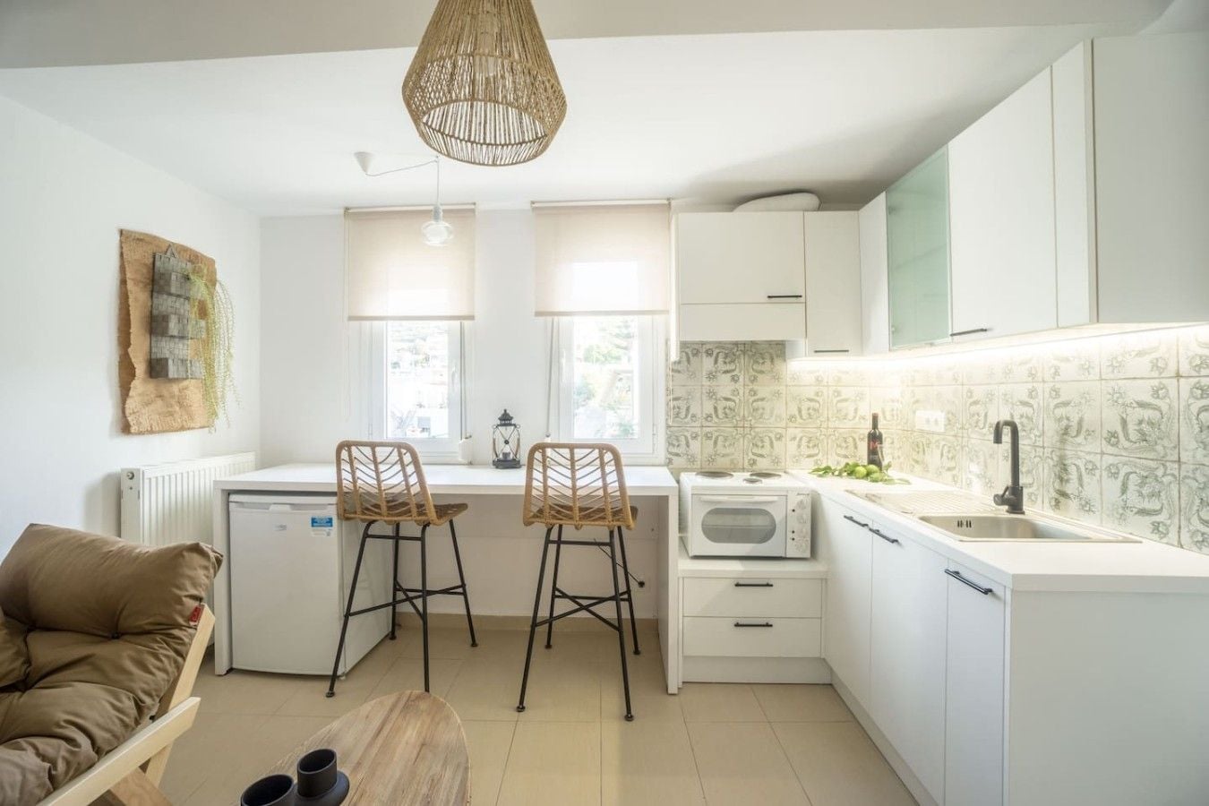 A compact kitchen with white cabinets, a built-in microwave, and a sink. There are two bar stools and a small dining table.