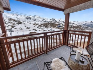 A balcony with a view of snow-covered mountains, a chair, a small table with a kettle, and a fur throw.