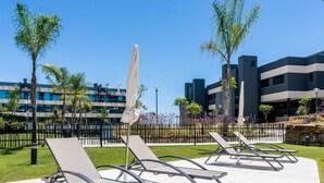 A modern outdoor seating area with sun loungers and umbrellas, surrounded by a well-maintained lawn and palm trees, with contemporary buildings in the background.