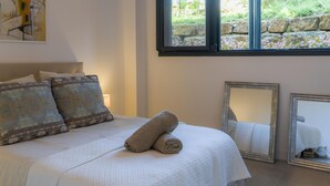 A neatly made bed with patterned pillows, a rolled towel, and a large window with a stone wall view.