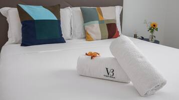 A neatly made bed with a white duvet, a rolled-up towel with a logo, and colorful pillows.