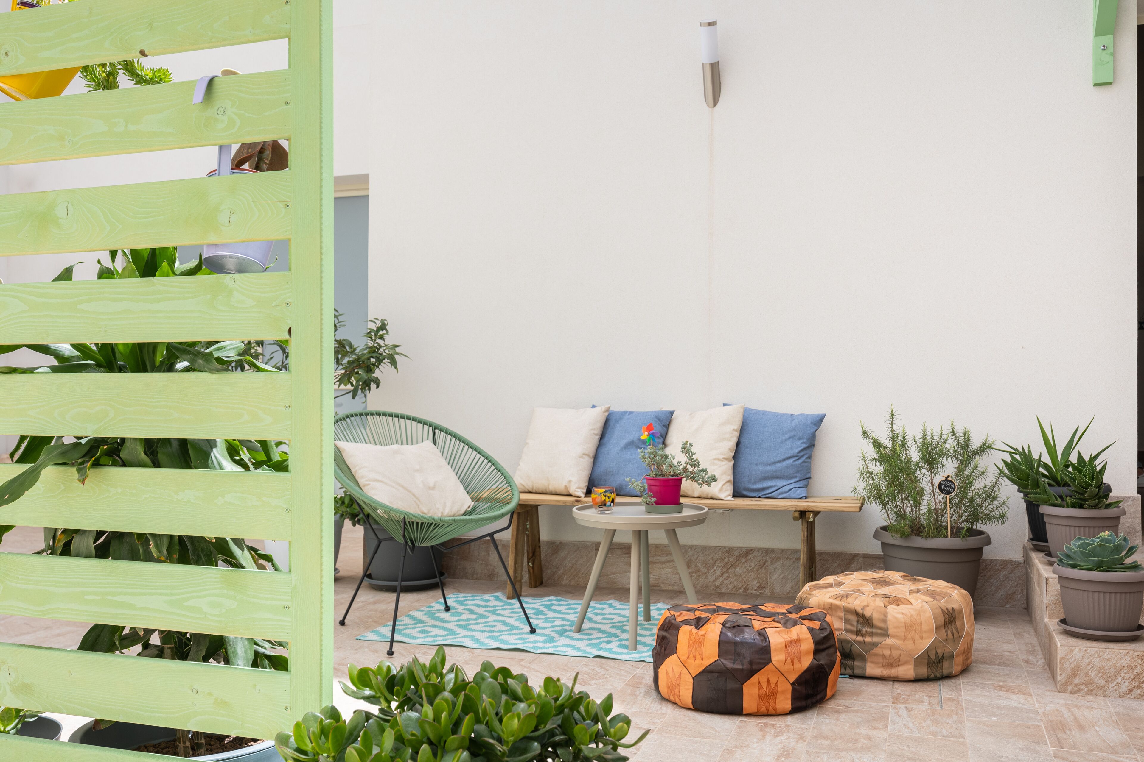 A cozy outdoor seating area with a wooden bench, a green chair, and a small table. There are several potted plants and a decorative orange cushion.