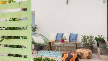 A cozy outdoor seating area with a wooden bench, a green chair, and a small table. There are several potted plants and a decorative orange cushion.