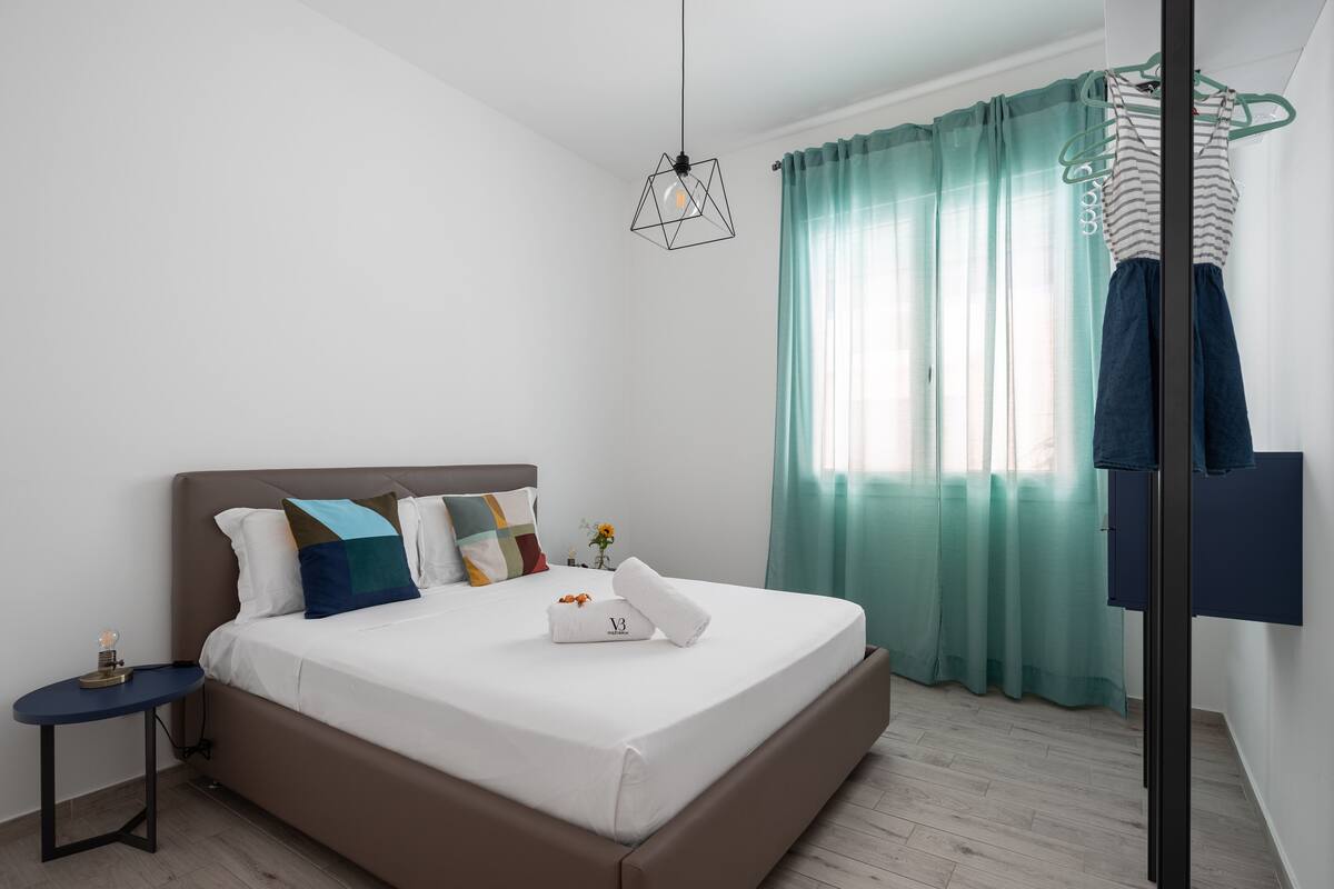 Une chambre moderne avec un grand lit, une table de chevet, un luminaire suspendu et une fenêtre avec des rideaux.