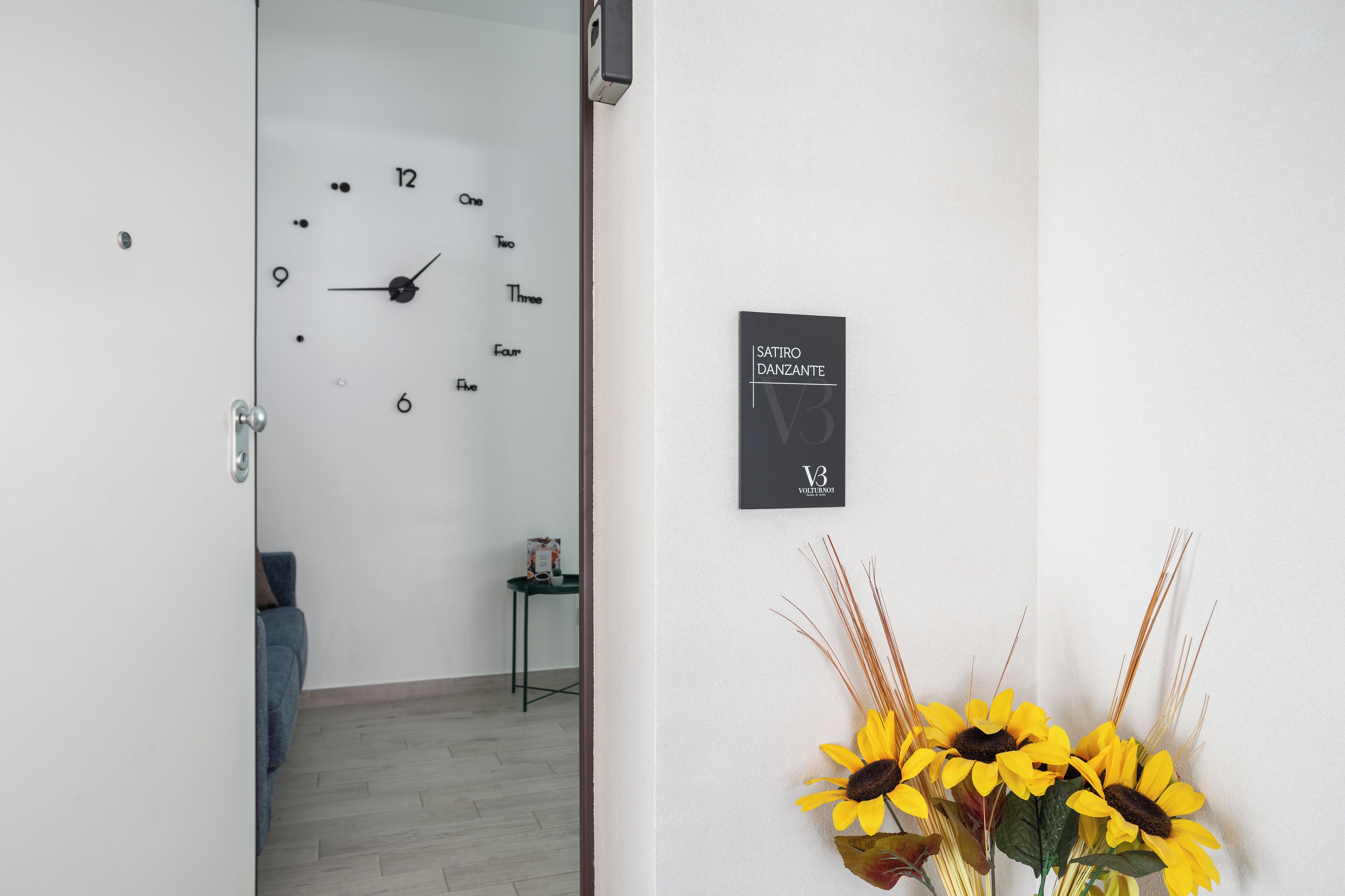 A room with a wall clock, a sign reading "SATIRO DANCANTE V3", and a vase with sunflowers.