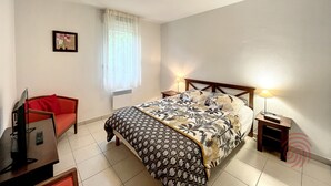 A bedroom with a bed, a TV, a chair, and a window with curtains. - Charming 1BR Apt with Large Terrace, A/C, WiFi, Near Town Center & Spa, Pet-Friendly! (Lamalou-les-Bains)