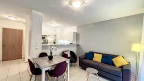 A modern living room with a dining area, a sofa with colorful cushions, and a small side table. - Charming 1BR Apt with Large Terrace, A/C, WiFi, Near Town Center & Spa, Pet-Friendly! (Lamalou-les-Bains)