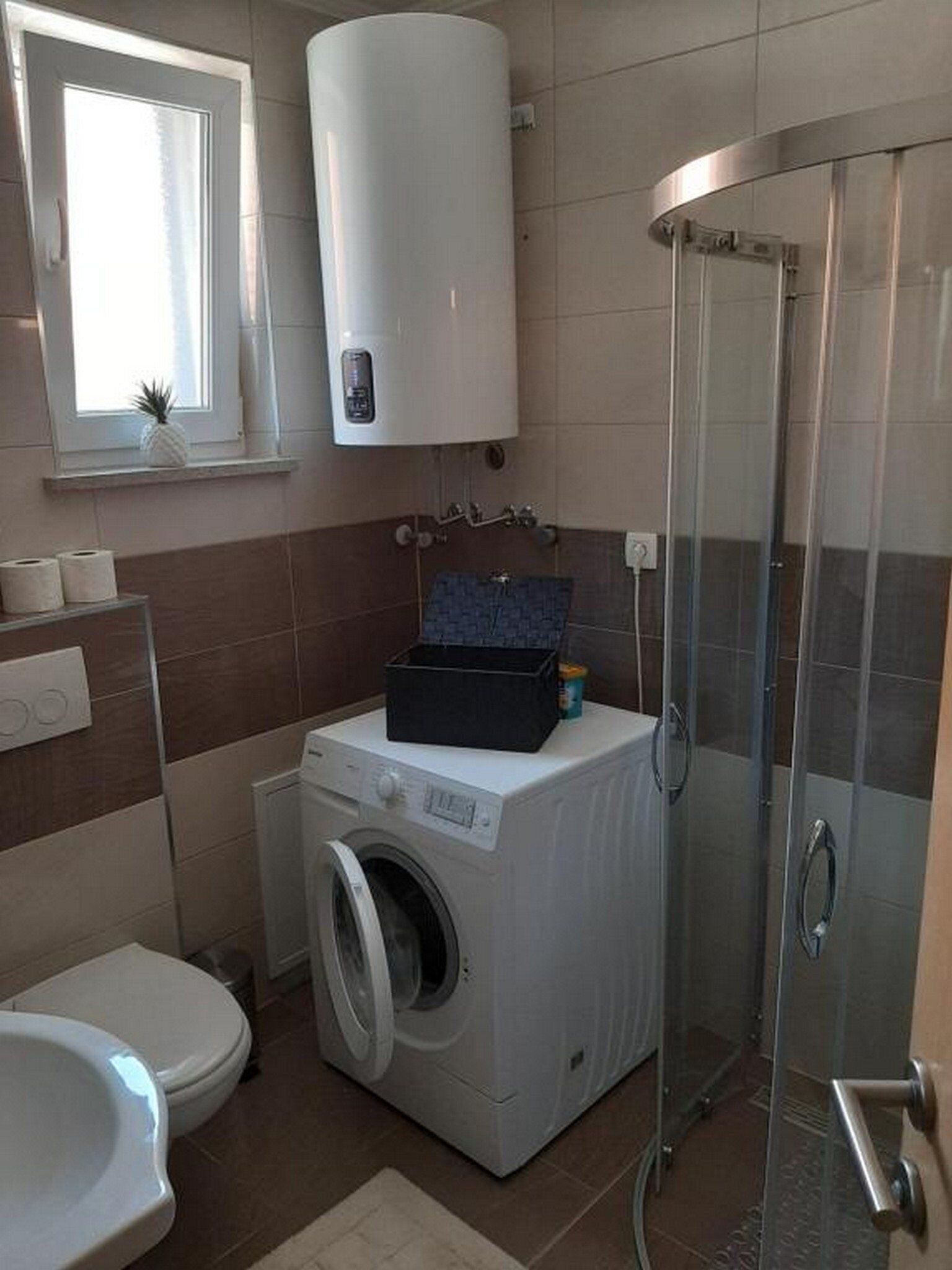A compact bathroom with a washing machine, a toilet, and a glass shower enclosure.