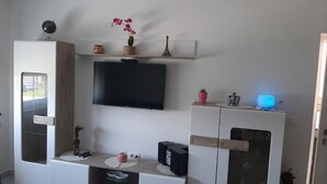 Un saló modern amb un televisor de pantalla plana muntat a la paret, un moble de fusta amb calaixos, una petita prestatgeria amb objectes decoratius i un aparell amb llums blaves sobre un moble.