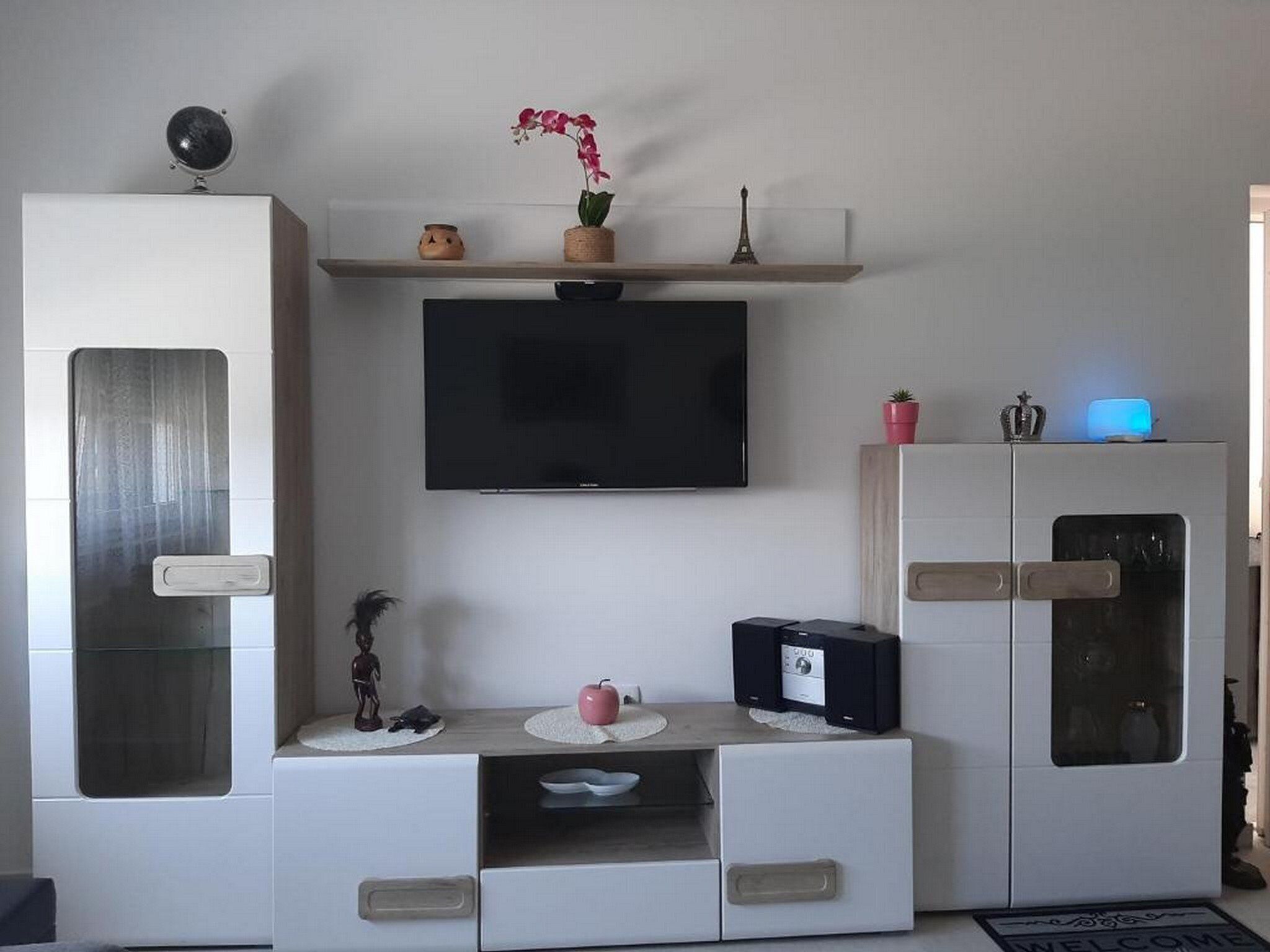 A modern living room with a flat-screen TV mounted on the wall, a small table with decorative items, and a cabinet with a built-in oven.
