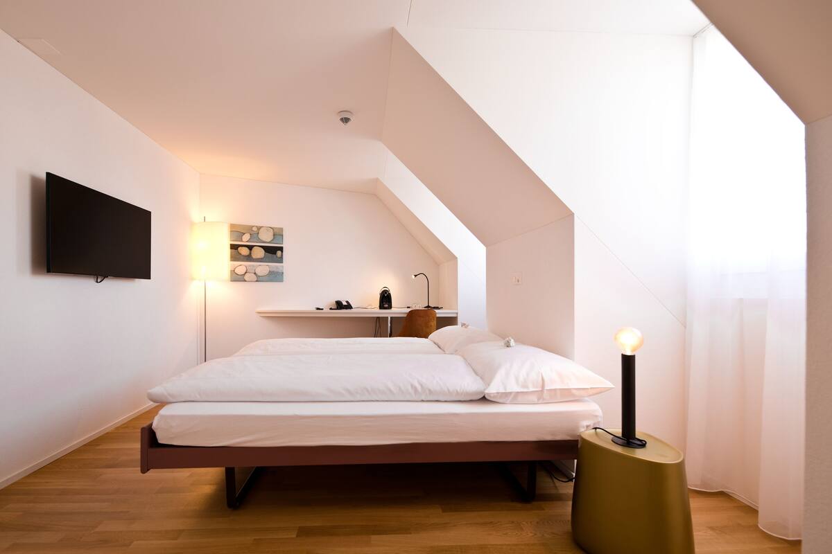 A bedroom with a bed, a bedside table with a lamp, a wall-mounted TV, and a ceiling light.