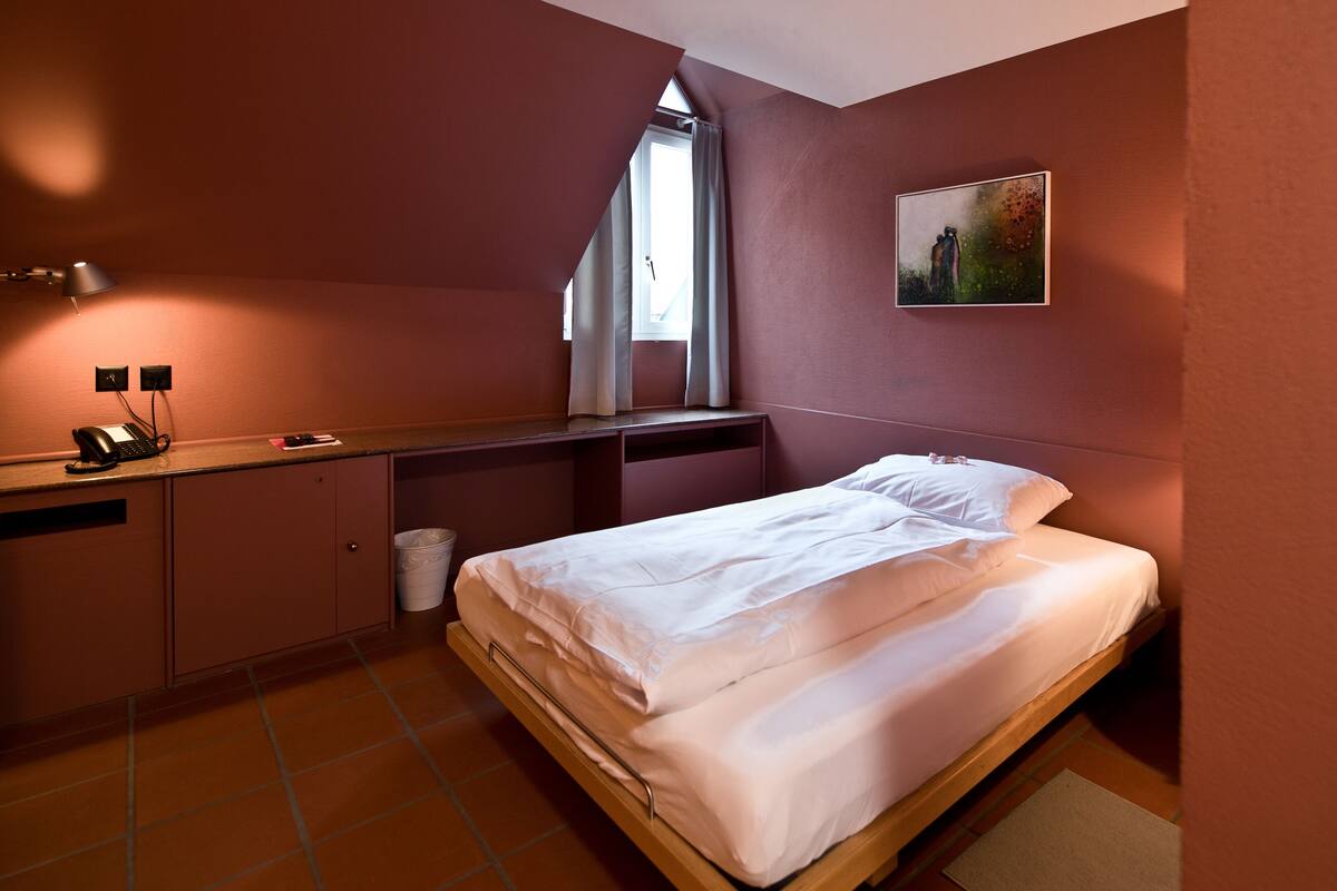 A bedroom with a bed, a desk, a chair, a lamp, a window, and a painting on the wall.