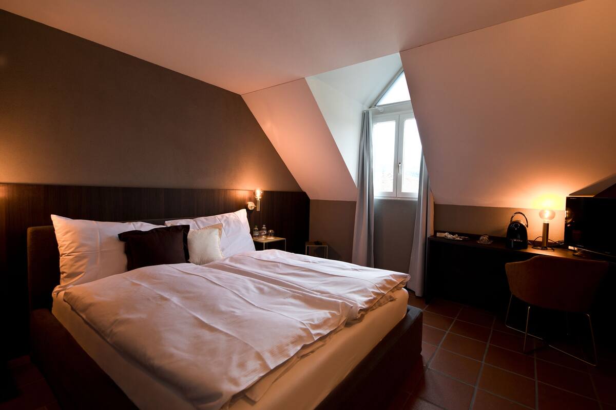 A hotel room with a large bed, a desk, and a chair. There is a window with curtains, a nightstand with a lamp, and a wall-mounted TV.