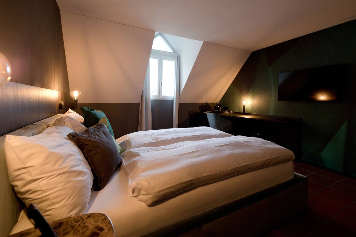 A bedroom with a bed, pillows, a nightstand, a TV, and a window.