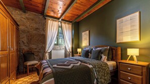 Ein Schlafzimmer mit Steinwand, Holzbalkendecke, einem Bett mit vielen Kissen, einem Nachttisch mit Lampe und einer Holzkommode.