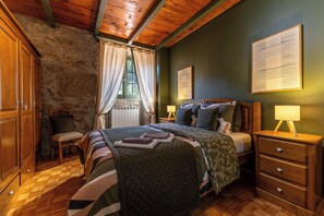 Un dormitorio con pared de piedra, vigas de madera en el techo, una cama con varios almohadones, una mesita de noche con lámpara y un aparador de madera.