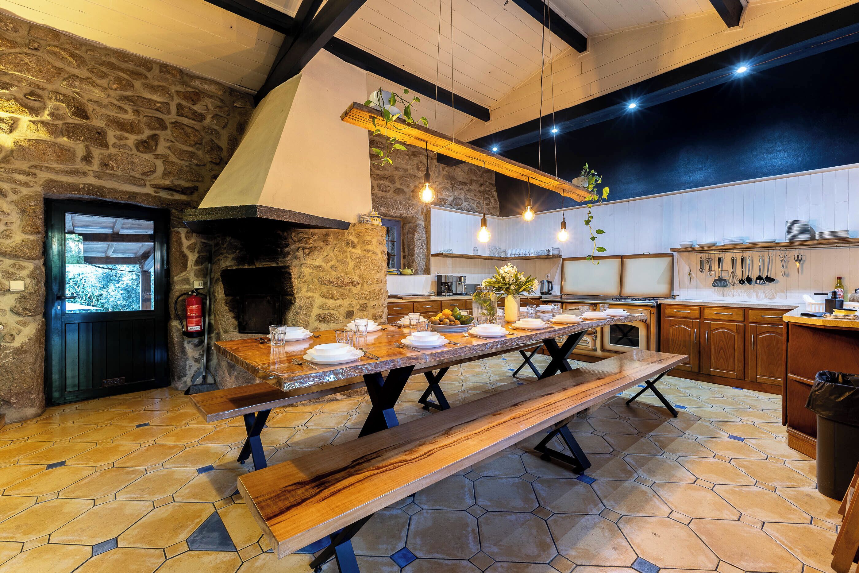 Une cuisine rustique avec un mur en pierre, des poutres en bois, une grande table en bois dressée pour le repas et un luminaire suspendu.