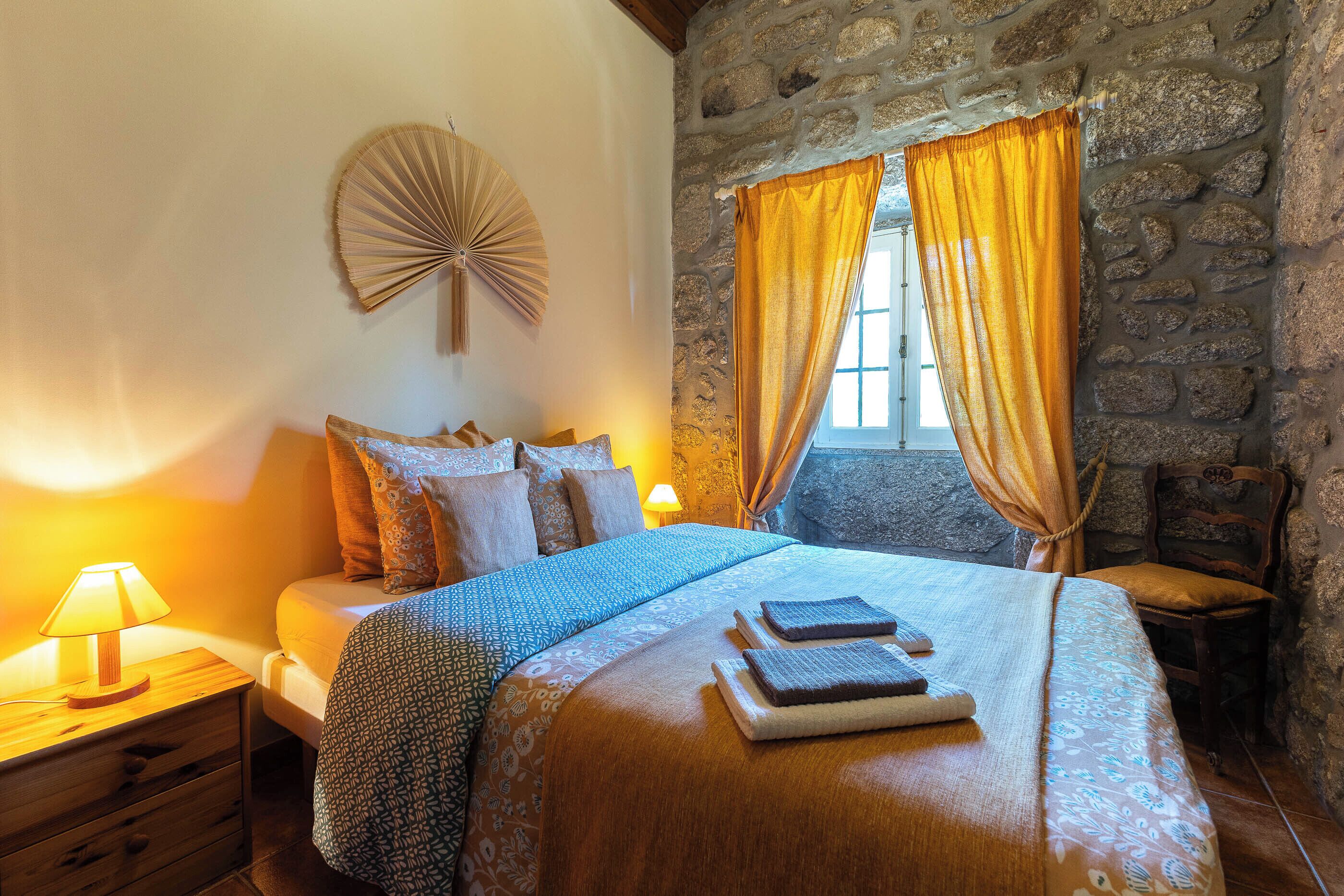 A bedroom with a stone wall, a bed with patterned bedding, a bedside lamp, and a window with curtains.