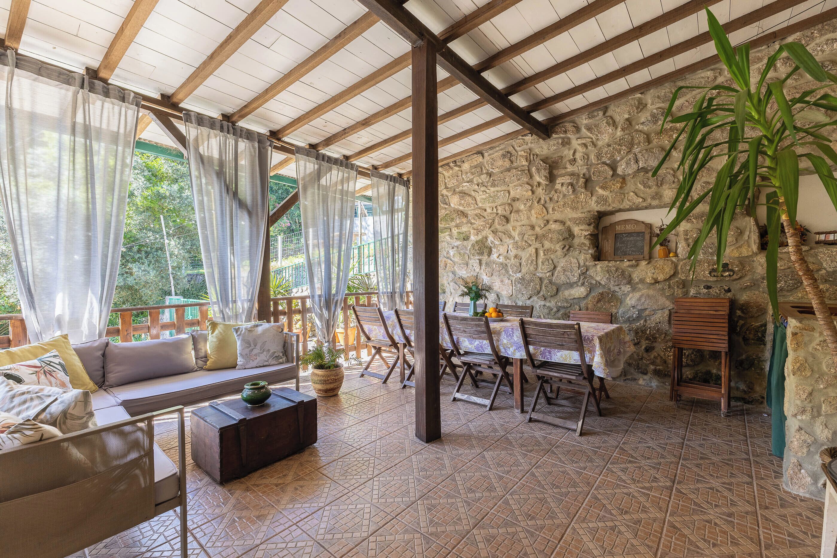A covered outdoor seating area with a stone wall, wooden furniture, and a table with chairs.