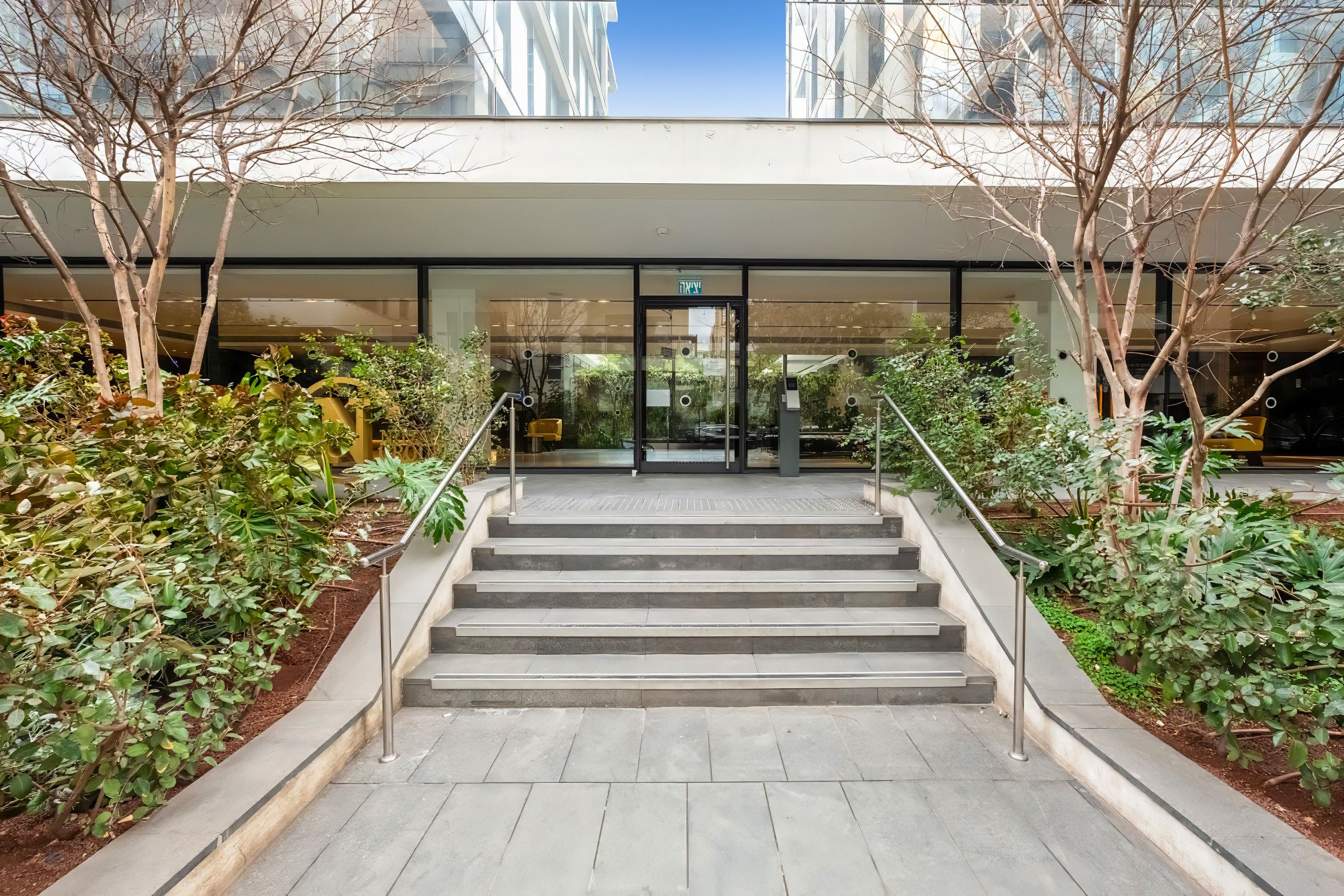 A modern building entrance with a glass door, a staircase, and surrounding greenery.