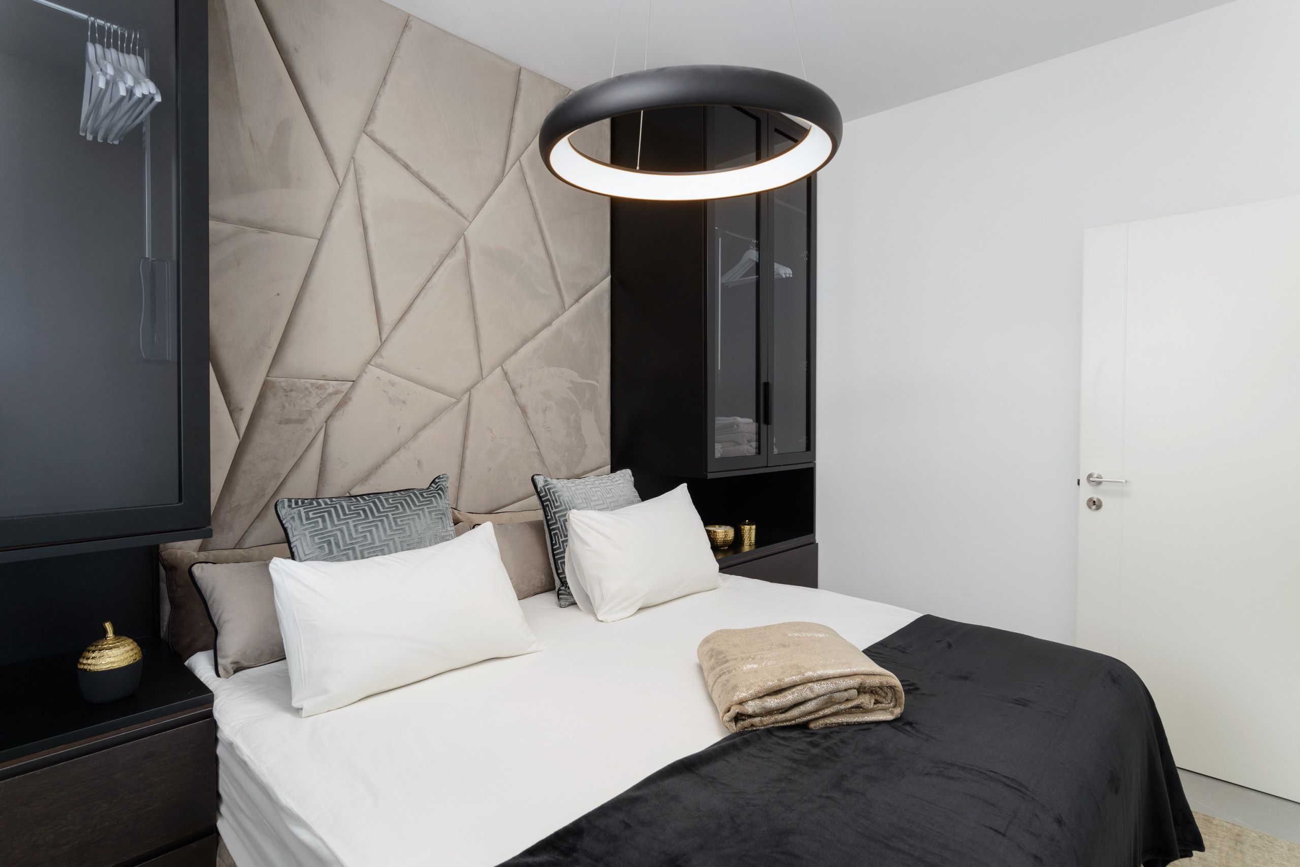 A modern bedroom with a large bed, a headboard with a geometric pattern, a bedside table with a lamp, and a wardrobe.