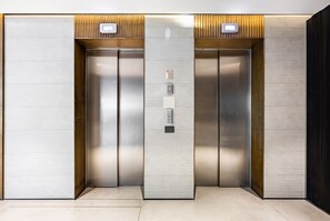 A modern elevator with metallic doors and control panels, featuring a textured wall panel and a light fixture above each door. - High End & City View by FeelHome (Tel Aviv)