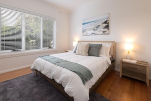Een slaapkamer met een bed, nachtkastje, lamp, schilderij aan de muur en raam met jaloezieën.