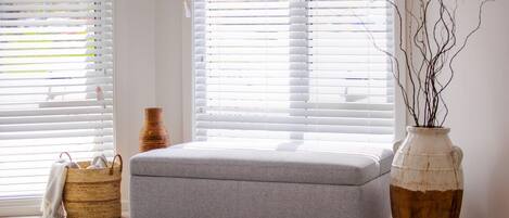 Una habitación con un reposapiés gris, una cesta tejida, un florero con ramas y una alfombra estampada.