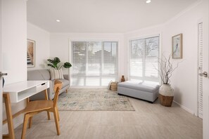 Een moderne woonkamer met een witte bank, een houten stoel, een bureau en een groot raam met jaloezieën.