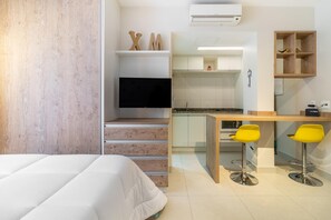 A modern hotel room with a bed, a TV, a kitchen area, and two yellow chairs.