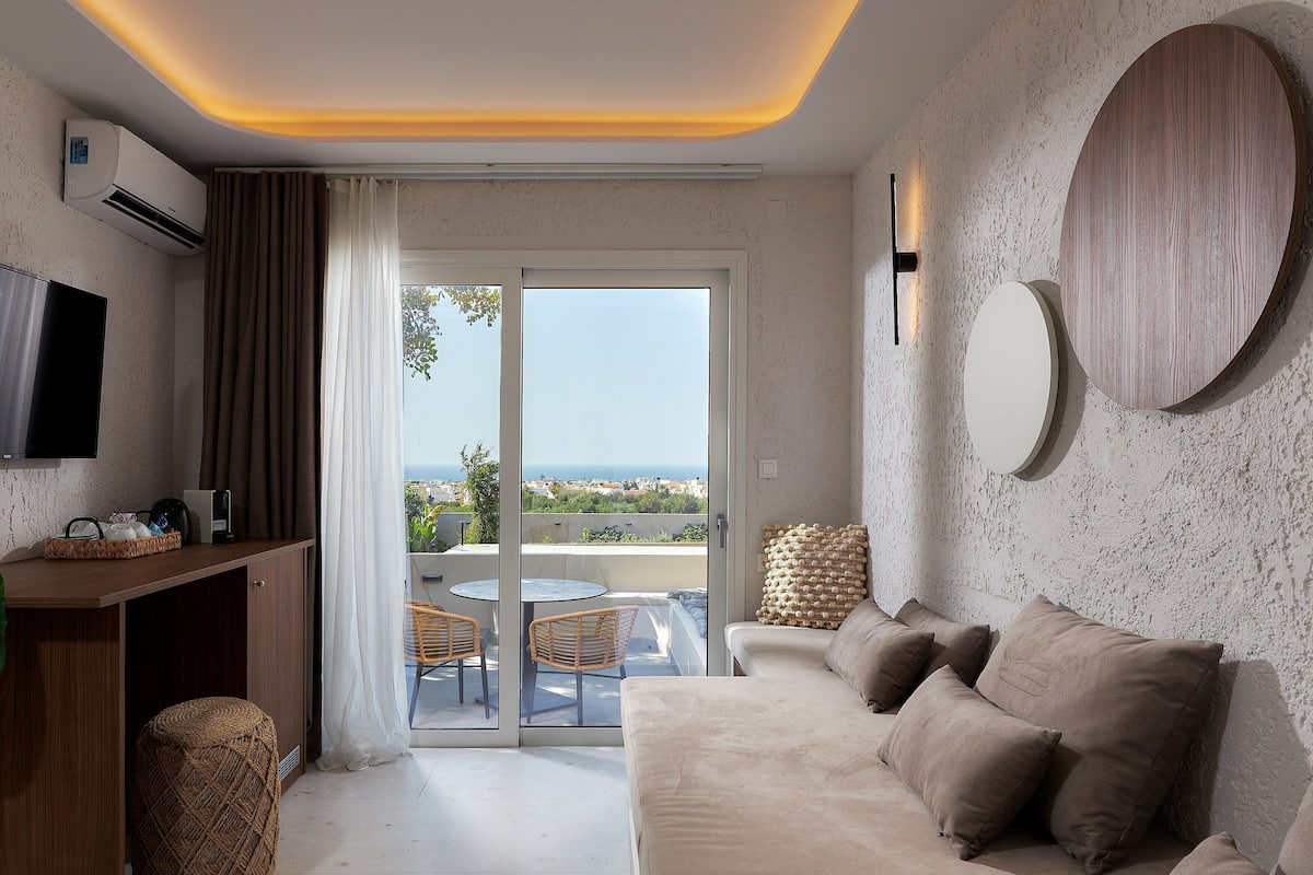 A modern room with a sofa, a TV, a dining area, and a view of the sea.