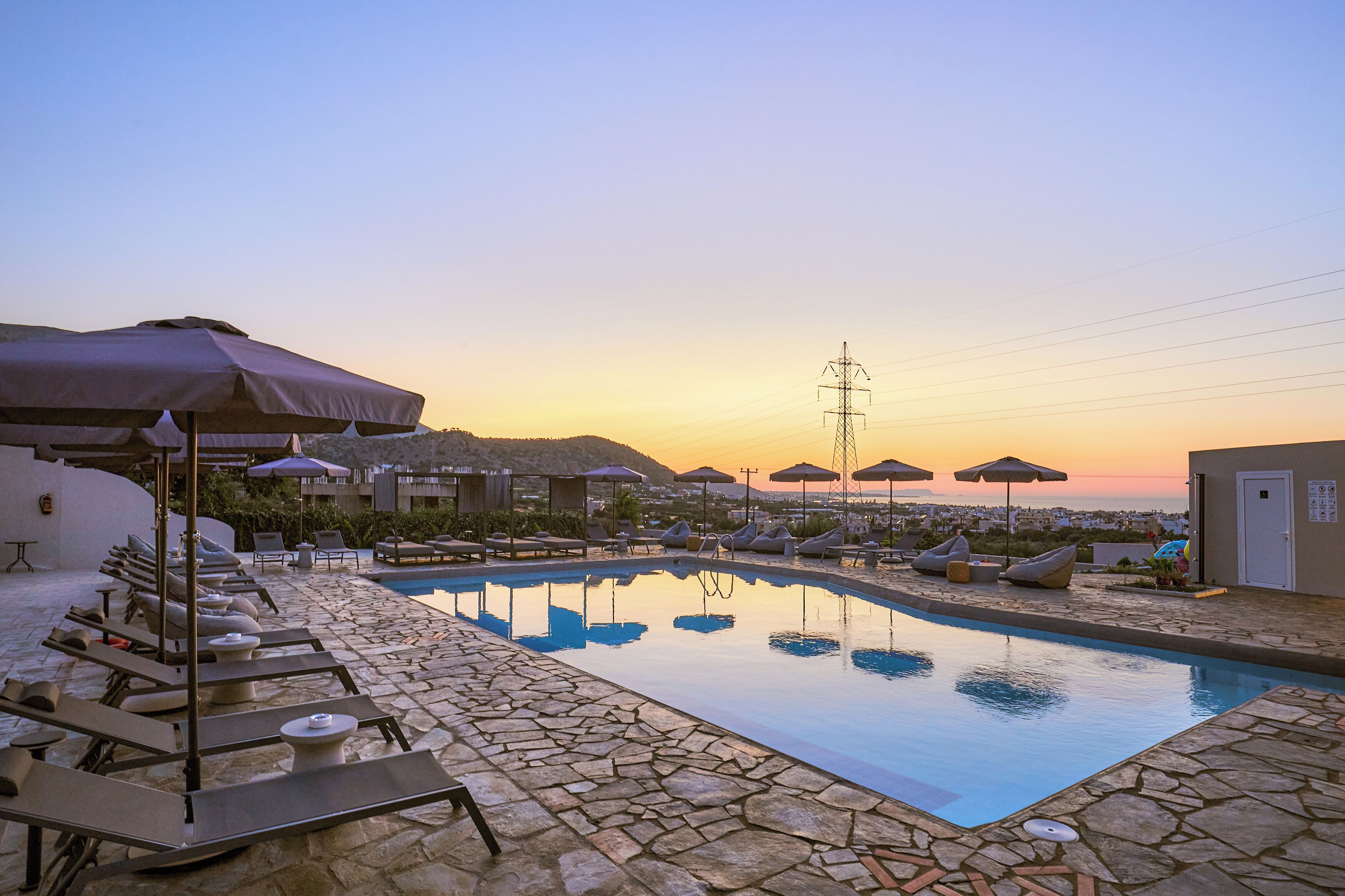 A poolside area with lounge chairs and umbrellas, a stone-tiled pool, and a view of the sea at sunset.