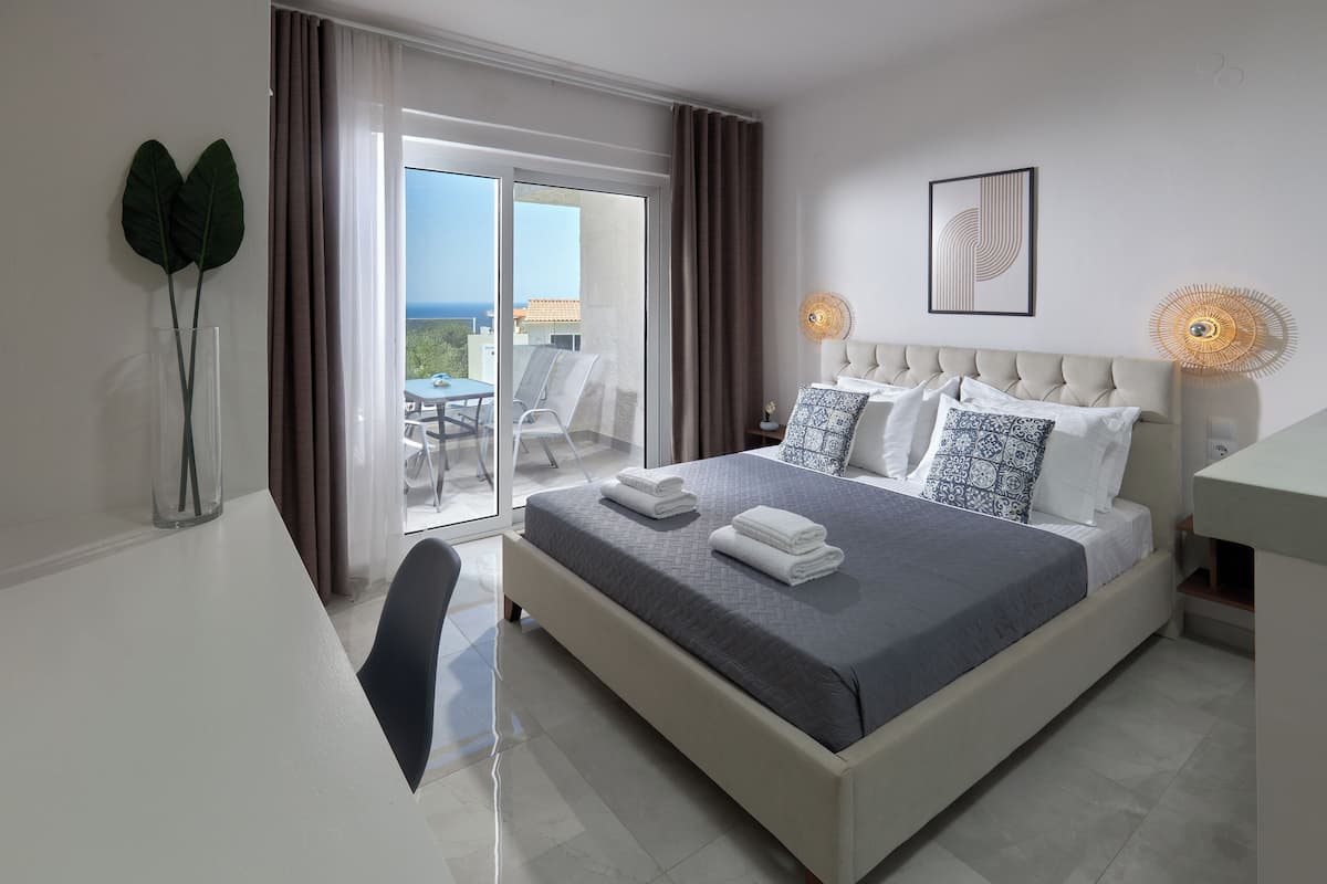 A modern hotel room with a large bed, a balcony with a table and chairs, and a view of the sea.