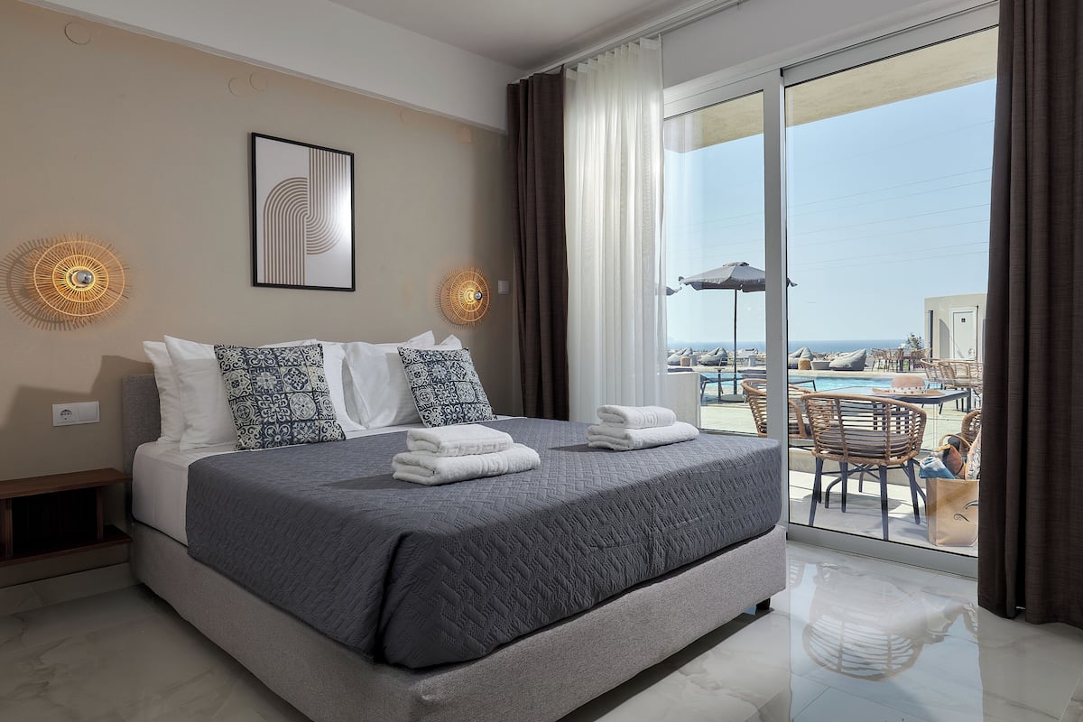A modern hotel room with a large bed, a view of the pool, and a balcony with outdoor furniture.