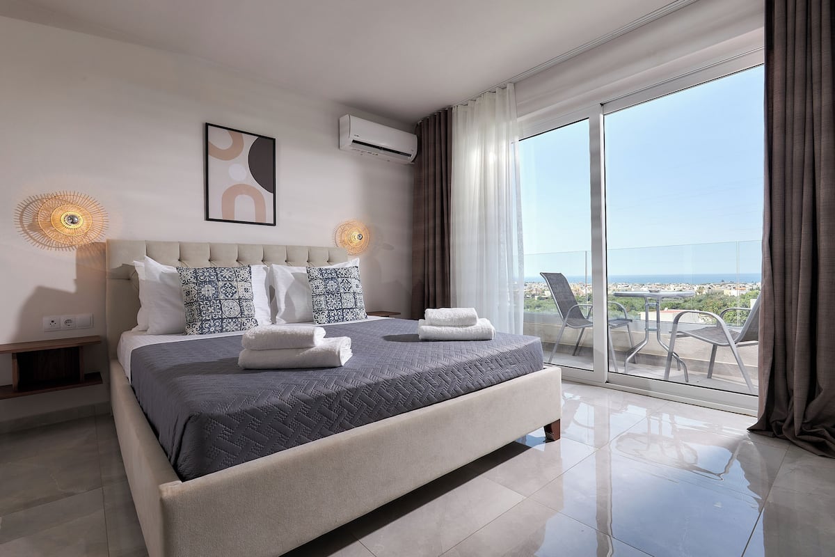 A modern hotel room with a large bed, a view of the city, and a balcony with chairs.