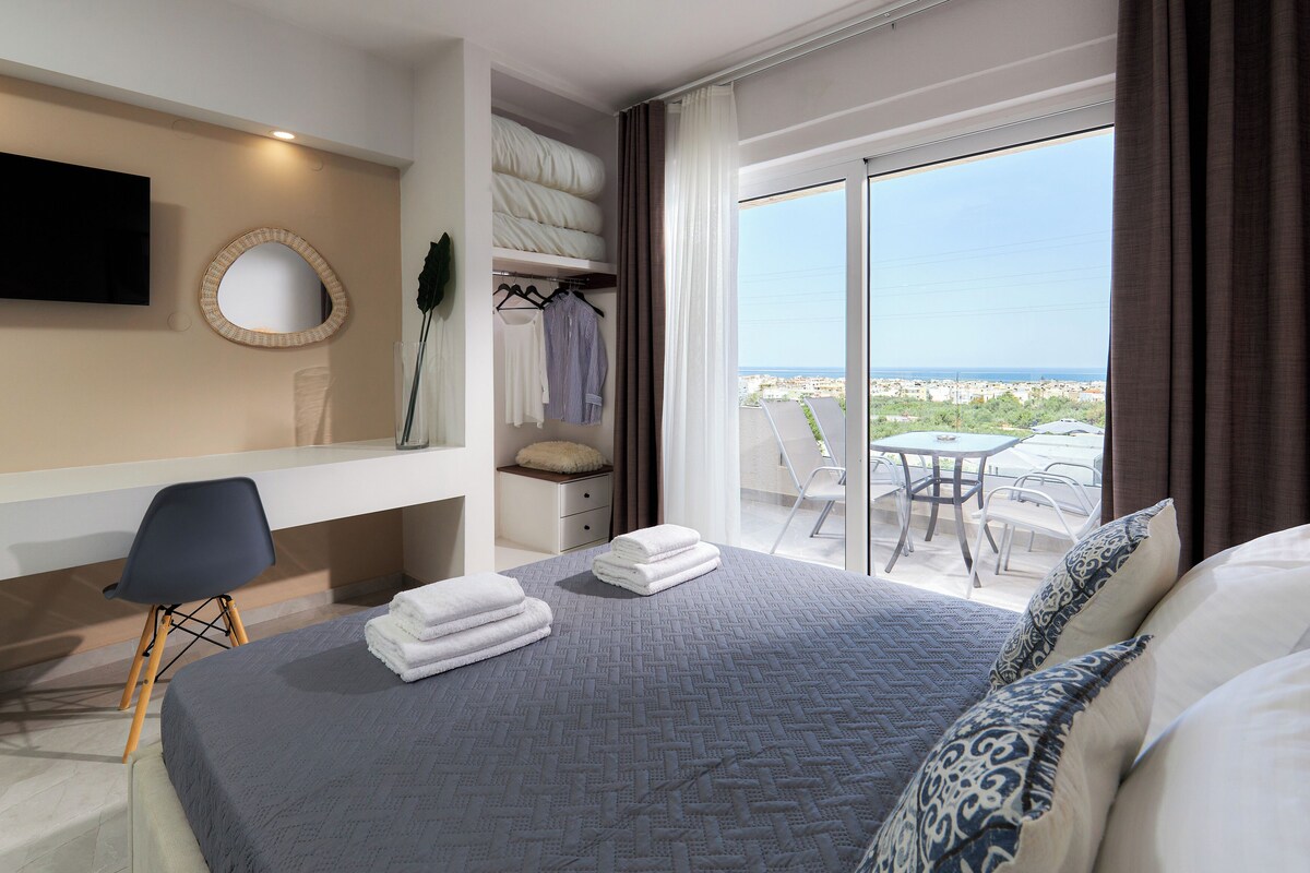 A hotel room with a bed, a desk, a chair, a TV, a balcony with a table and chairs, and a view of the city.
