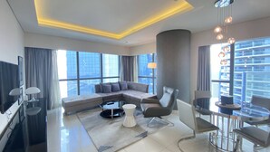 Un salon moderne avec un canapé d’angle, une table basse en verre, une table de salle à manger ronde avec des chaises, un téléviseur et une grande fenêtre donnant sur la ville.