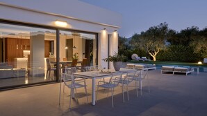 A modern outdoor dining area with a glass-enclosed kitchen, a pool, and a well-maintained garden.