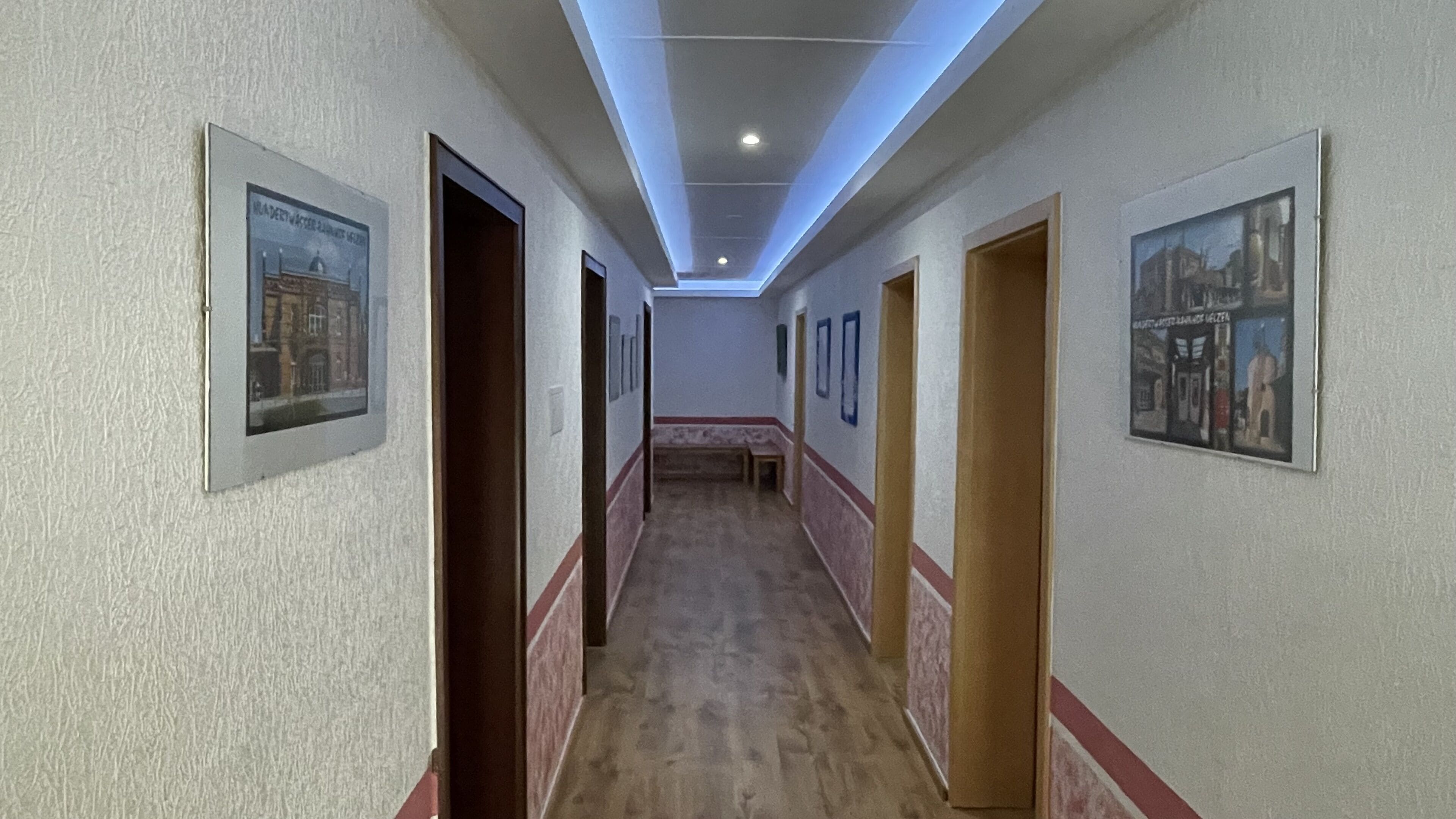 Un couloir avec des tableaux encadrés accrochés aux murs, un banc en bois et des portes donnant sur différentes pièces.
