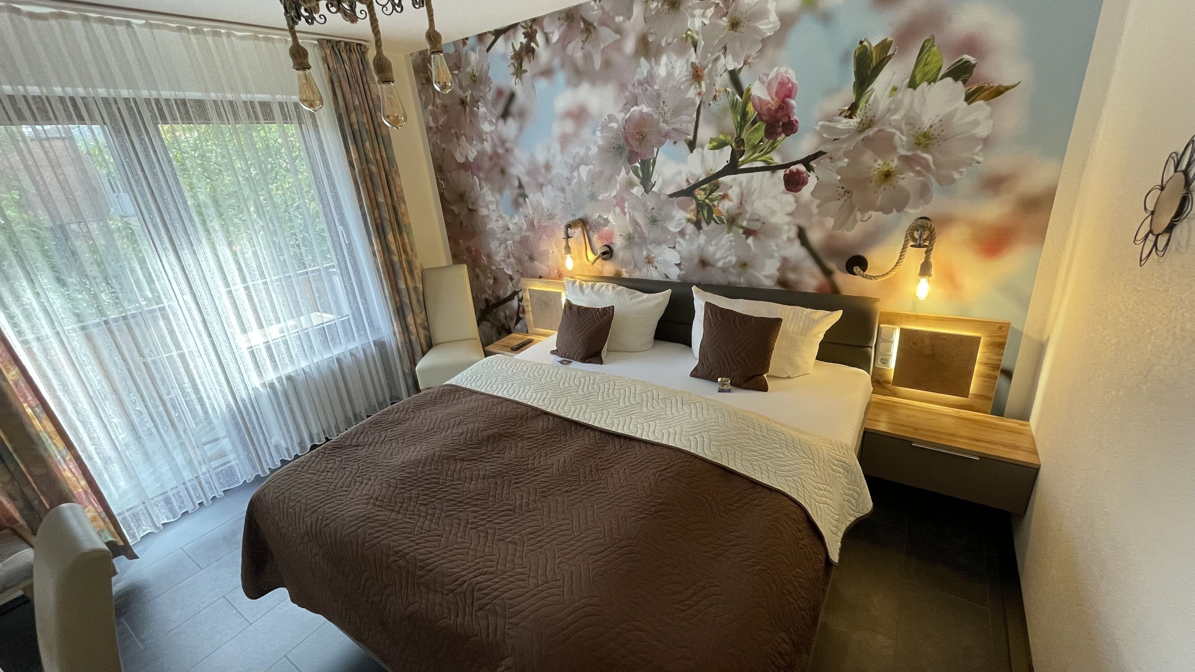 A hotel room with a large bed, a nightstand with a lamp, a wall-mounted light, and a view of cherry blossoms through the window.