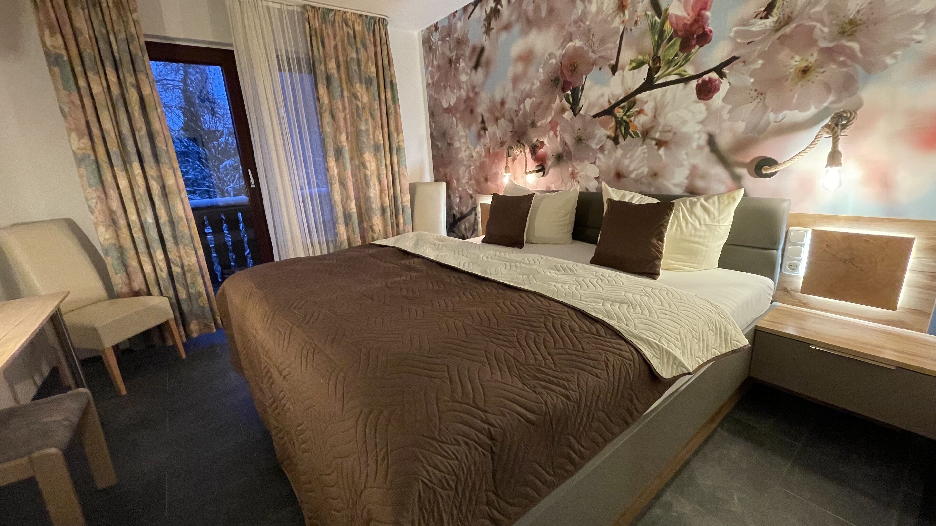 A hotel room with a large bed, a desk, and a chair. There is a window with curtains, a wall with floral wallpaper, and a nightstand with a lamp.