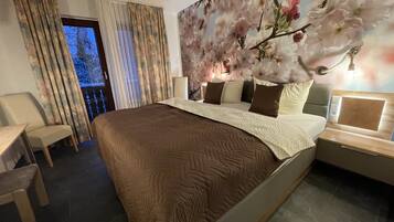 A hotel room with a large bed, a desk, and a chair. There is a window with curtains, a wall with floral wallpaper, and a nightstand with a lamp.