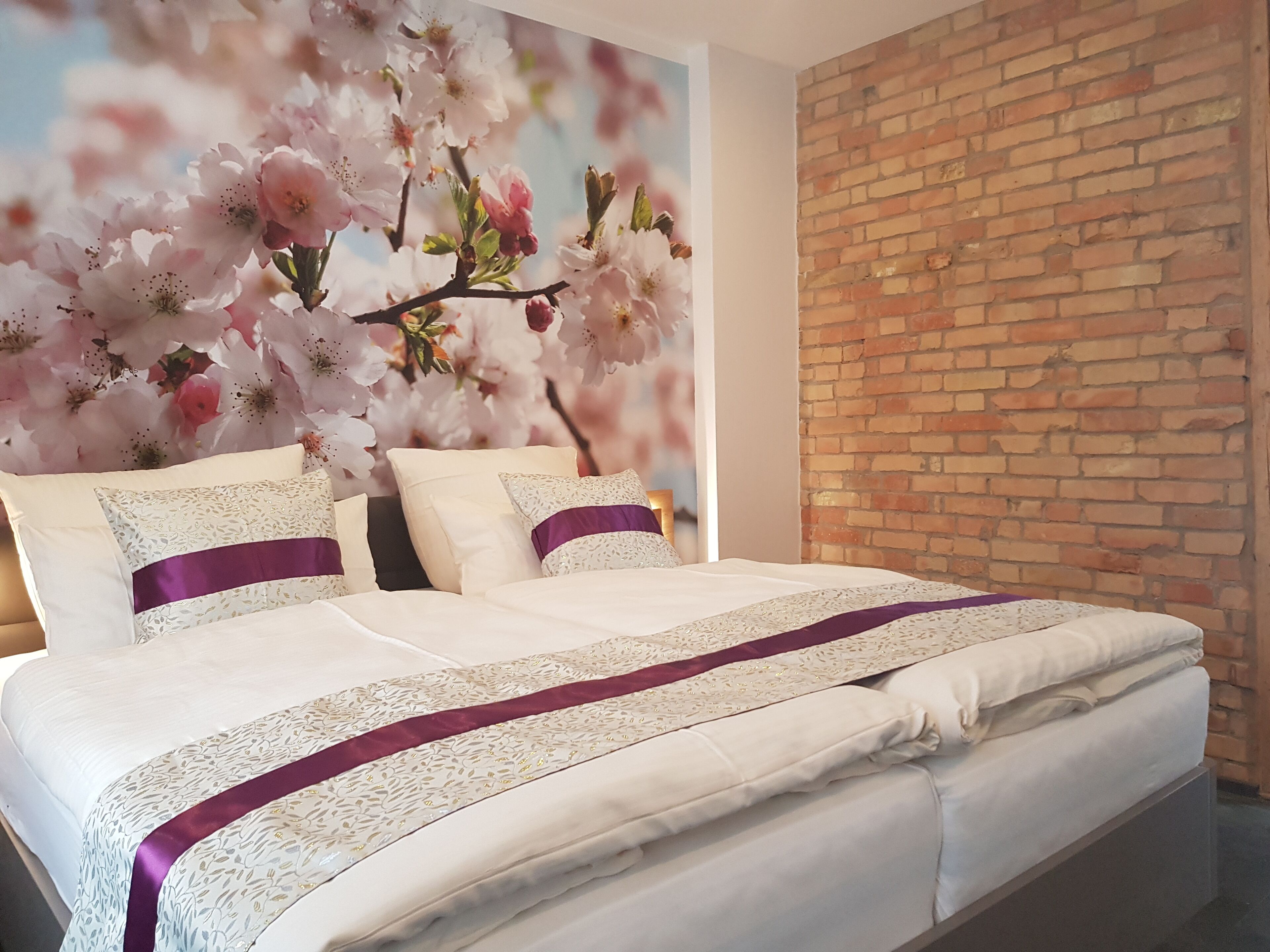 A bedroom with a brick wall, a bed with white bedding and purple accents, and a floral mural on the wall.
