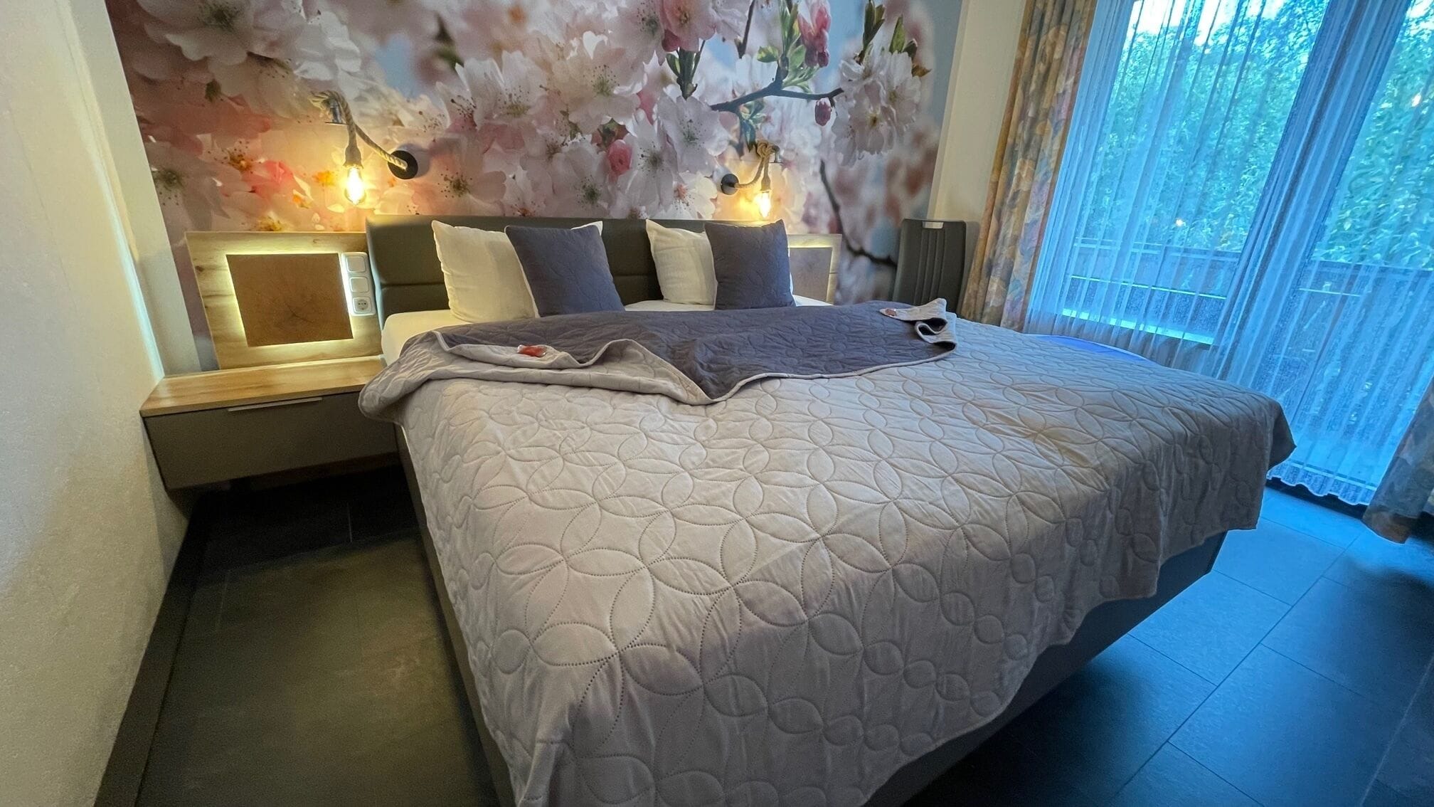 Ein ordentlich bezogenes Bett mit gesteppter Decke, ein Nachttisch mit Lampe und eine Blumenwandmalerei.