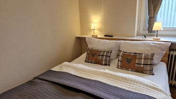 Un lit bien fait, recouvert d’une couverture marron matelassée, avec deux oreillers ornés de motifs en forme de cœur et une table de chevet sur laquelle se trouve une lampe.