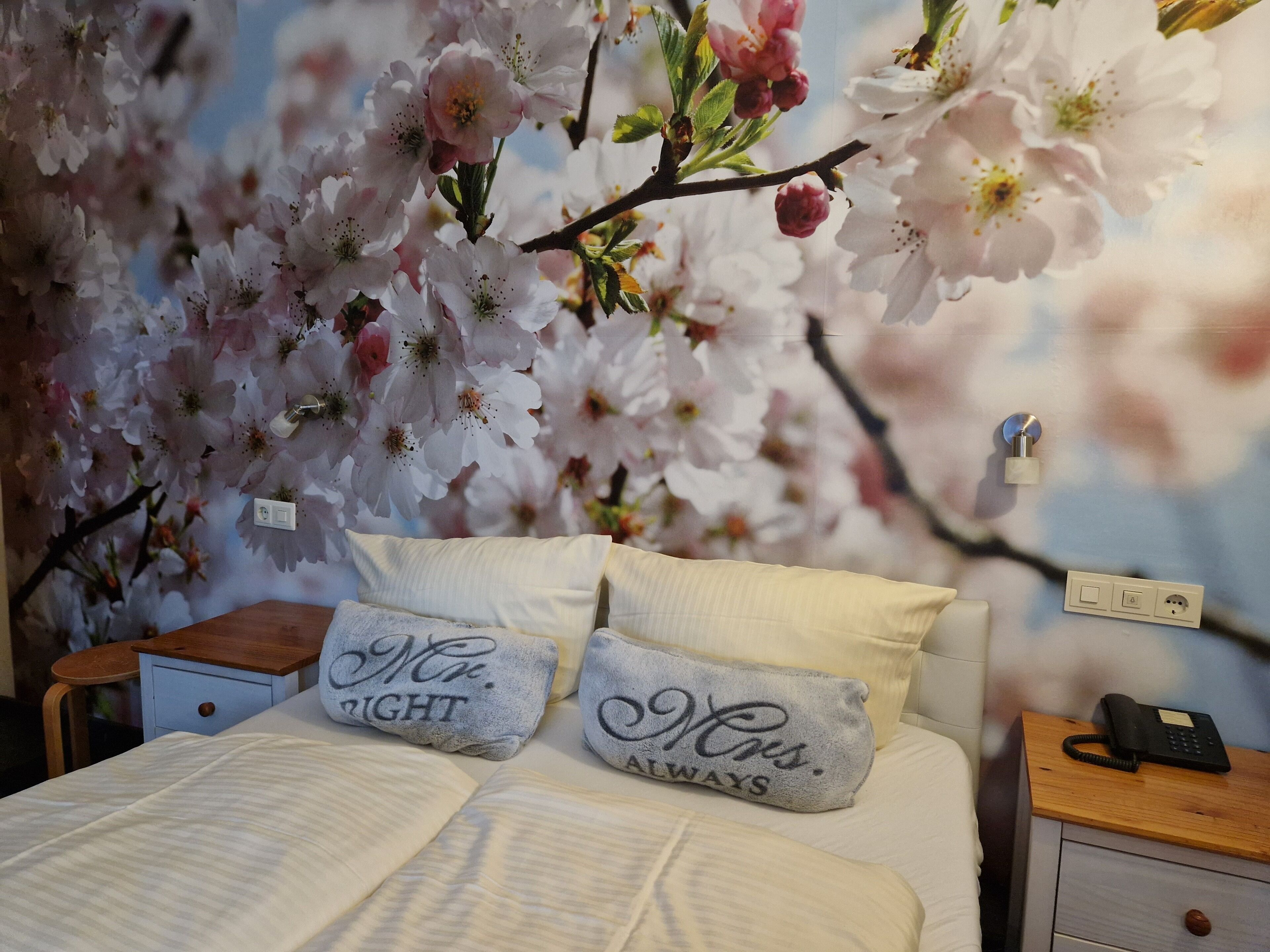 Camera da letto con un letto, due cuscini, un comodino, un telefono fissato al muro e un murale con fiori di ciliegio.