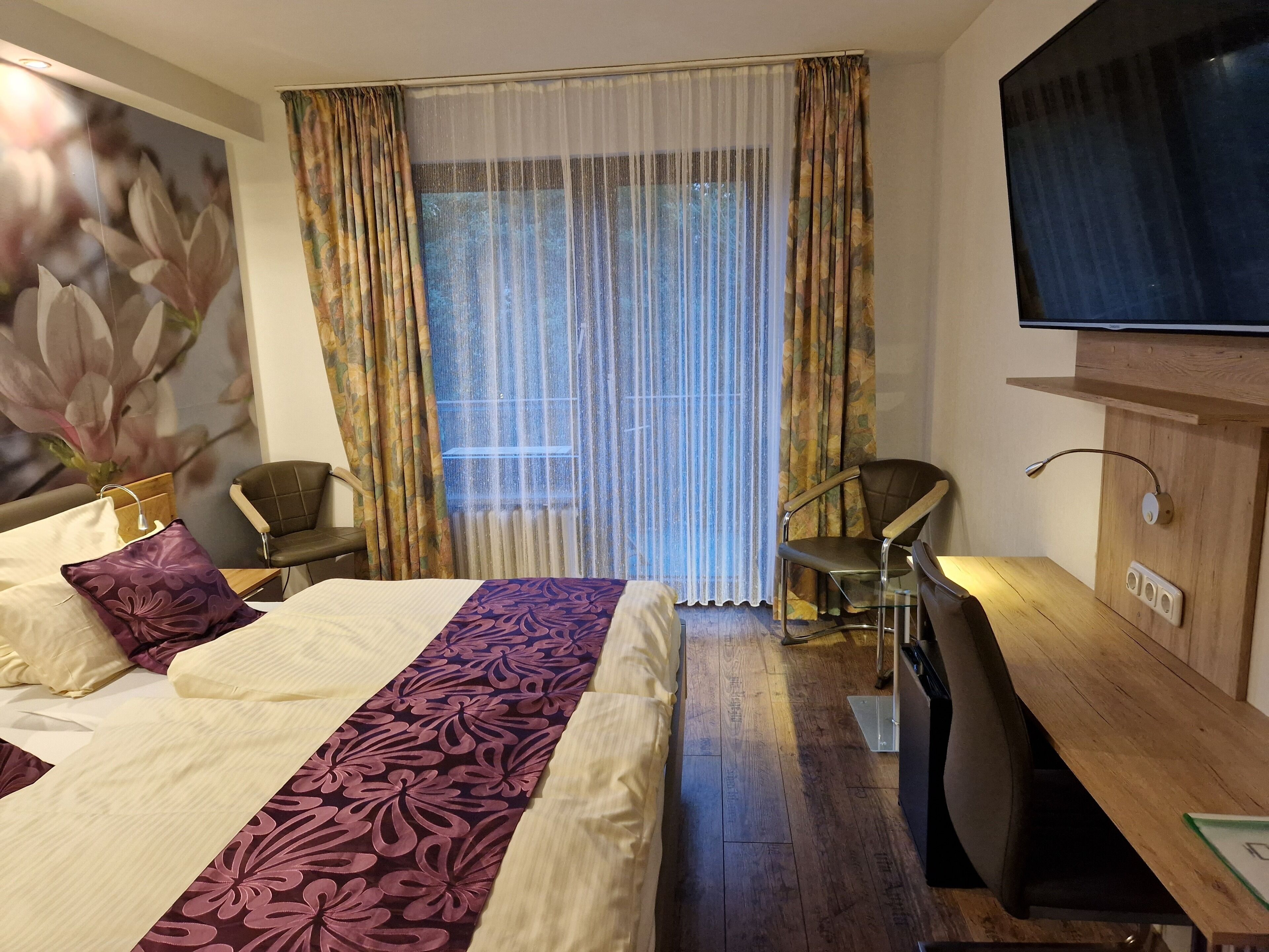 A hotel room with a bed, two chairs, a desk, a television, and a window with curtains.