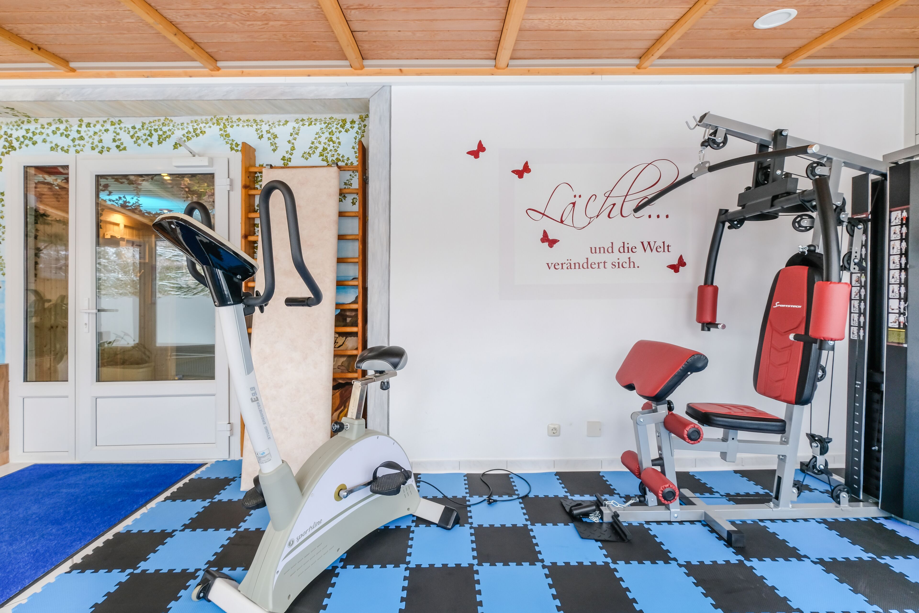 A home gym with exercise equipment, a blue and black mat floor, and a motivational wall quote.