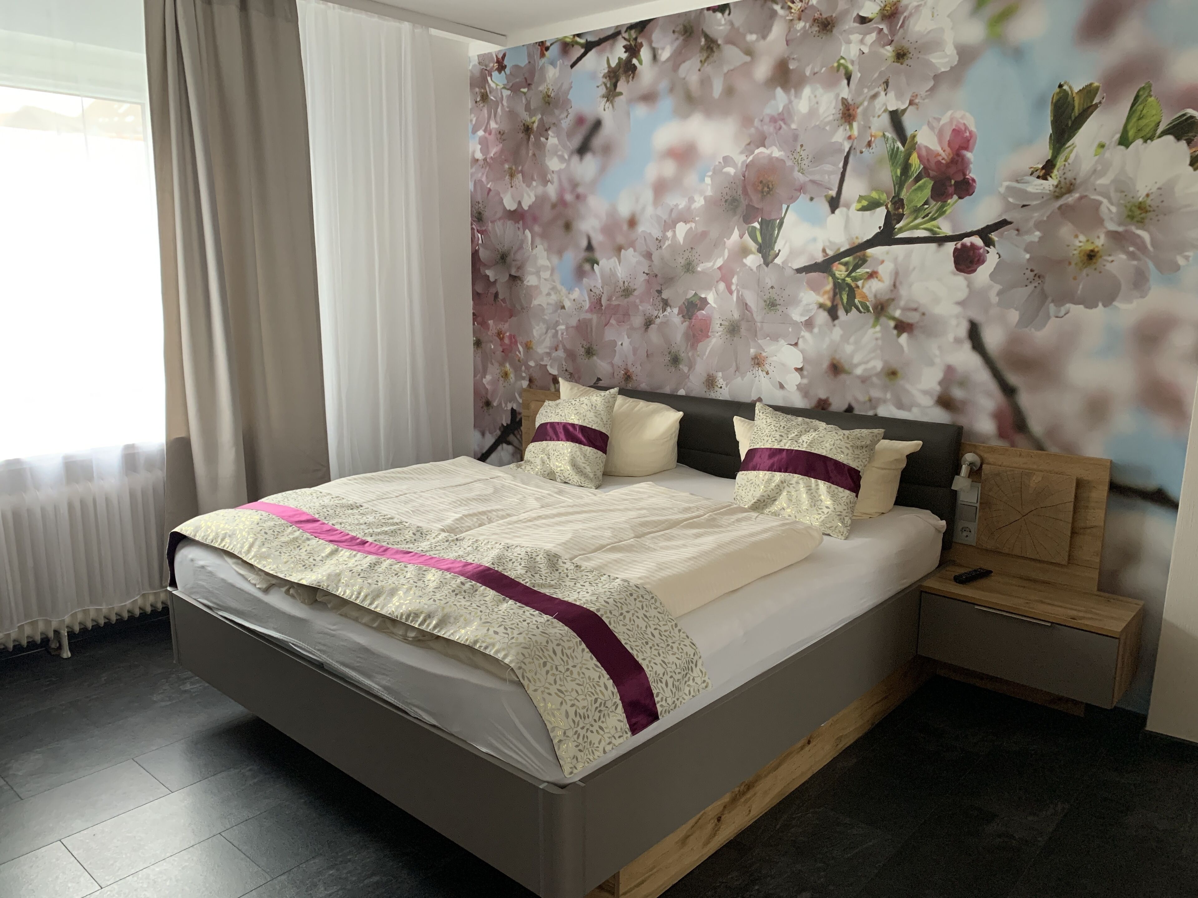 A bedroom with a large bed, a nightstand, a wooden headboard, and a wall mural of cherry blossoms.