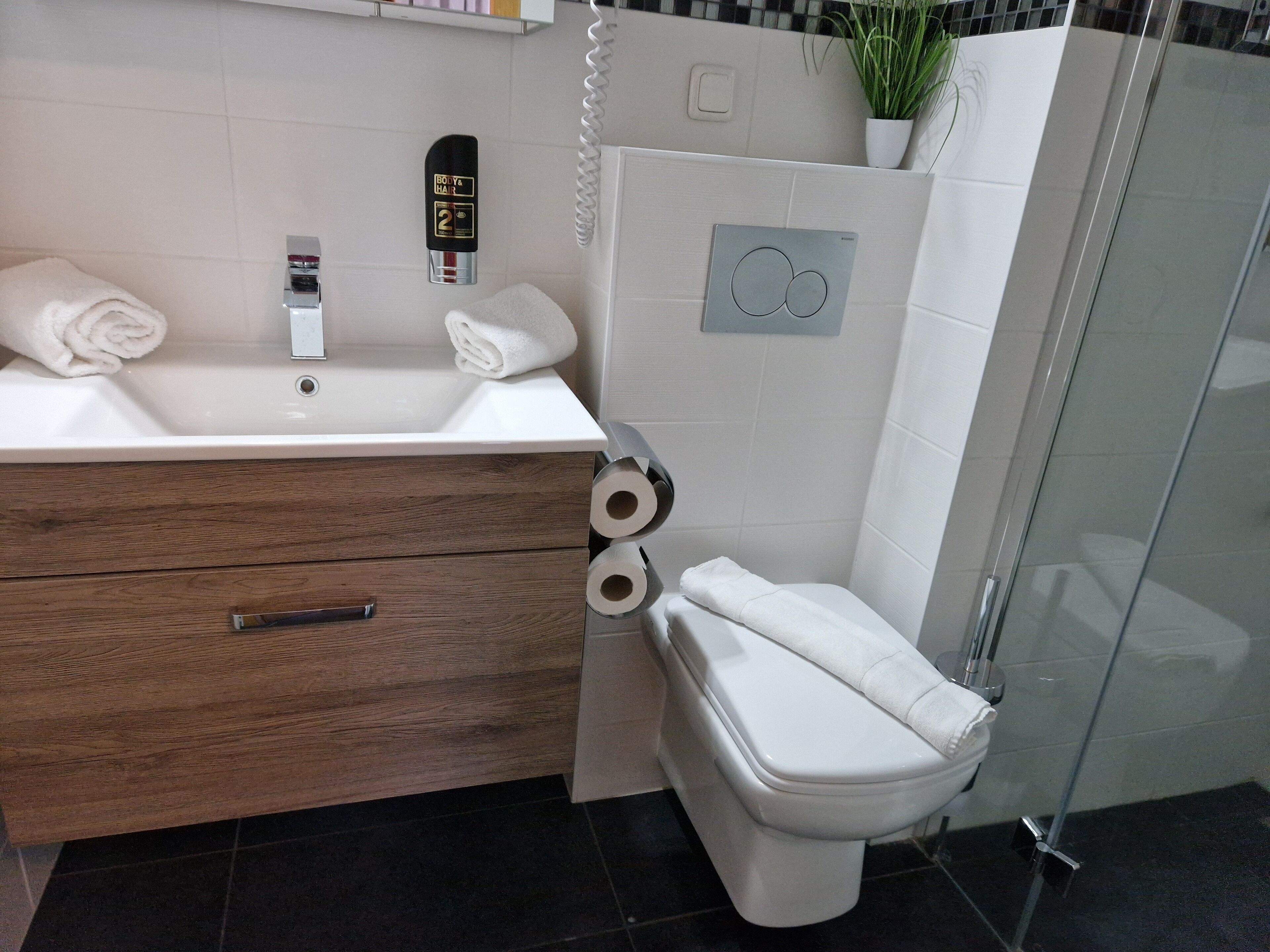 Un baño moderno con tocador de madera, lavamanos blanco y retrete. Hay dos toallas sobre el tocador y un dispensador de papel higiénico.